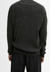 Personne portant un pull en maille côtelée gris foncé et un pantalon noir, vue de dos sur un fond clair uni.