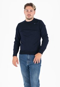 Temno modra jopica, okrogla izrezana vrata, žametni manšeti in rob, mehkega pletenega materiala, z logotipom na prsih, oblečena z svetlo modrimi kavbojkami.