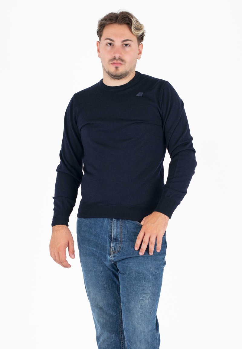 Temno modra jopica, okrogla izrezana vrata, žametni manšeti in rob, mehkega pletenega materiala, z logotipom na prsih, oblečena z svetlo modrimi kavbojkami.