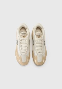 Beige nät- och lädersneakers med en krämfärgad tåskydd, snörningar och metalliska detaljer. Har en rundad form och texturerade inslag.