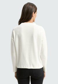 Pull blanc à texture avec une coupe décontractée, un col rond et des manches longues, associé à un jean noir. Vue arrière montrée.