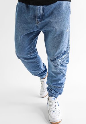 Light blue denim jogger pants with elastic cuffs, featuring side pockets and contrasting white logo detail. Paired with white sneakers.