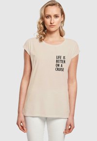 Helle beige T-Shirt mit aufgerollten Ärmeln und rundem Ausschnitt, mit schwarzem Text: "DAS LEBEN IST BESSER AUF EINER KREUZFAHRT." Glatte Stofftextur.