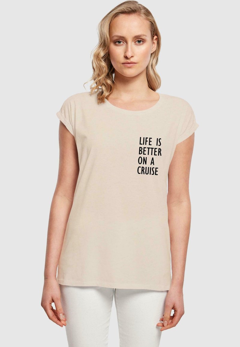 Helle beige T-Shirt mit aufgerollten Ärmeln und rundem Ausschnitt, mit schwarzem Text: "DAS LEBEN IST BESSER AUF EINER KREUZFAHRT." Glatte Stofftextur.