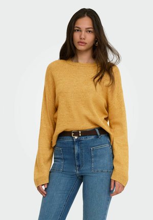 Femme portant un pull jaune moutarde rentré dans un jean bleu taille haute avec des poches plaquées à l'avant et une ceinture marron foncé.