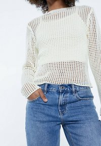 Maglione bianco lavorato a maglia con design a trama aperta e maniche lunghe, abbinato a jeans blu a vita alta con tasche frontali.