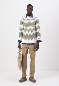 Strickpullover mit einem geometrischen Muster in Grün, Weiß und Braun. Kombiniert mit khaki-farbenen Hosen und braunen Schuhen. Hält eine hellbraune Tasche.