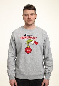 Sudadera gris con el texto "�¡Feliz Grinchmas!", una ilustración de la mano del Grinch sosteniendo un adorno navideño y acentos rojos.