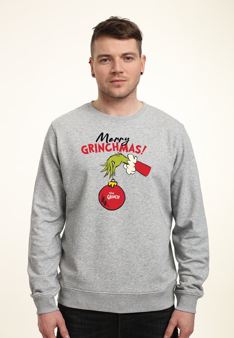 Sudadera gris con el texto "¡Feliz Grinchmas!", una ilustración de la mano del Grinch sosteniendo un adorno navideño y acentos rojos.