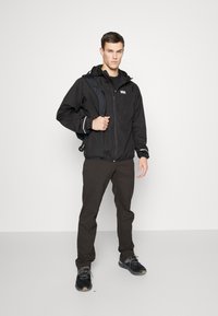 Crna vodootporna jakna s kapuljačom, s bijelim naglascima i logotipom, u kombinaciji s tamnim hlačama i crnim cipelama, nošena s crnim ruksakom.