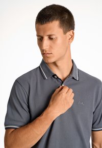 Szary polo z teksturowanej tkaniny, z kołnierzykiem na dwa guziki i białymi akcentami na rękawach. Wyszywane logo na klatce piersiowej. Zbliżenie na modela.