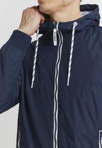 Chaqueta con capucha en color navy, hecha de un material suave y liviano, con cremallera frontal, cuerdas blancas en contraste y puños de canalé.