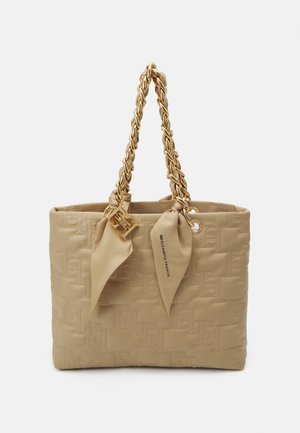 Bolso tote beige acolchado con asas de cadena dorada, pañuelos beige anudados y un colgante con el logo dorado sobre un fondo liso.
