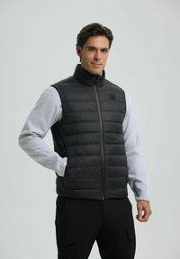 Een man in een zwart gewatteerd vest over een lichtgrijze longsleeve shirt met zwarte broek, staande met één hand in zijn zak tegen een eenvoudige achtergrond.