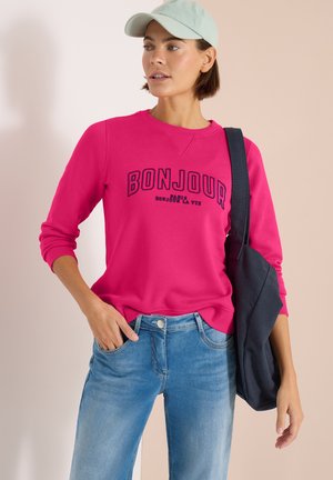 Femme portant une casquette vert clair, un sweat-shirt rose vif avec le texte « BONJOUR », un jean bleu et un sac à bandoulière noir sur un fond neutre.