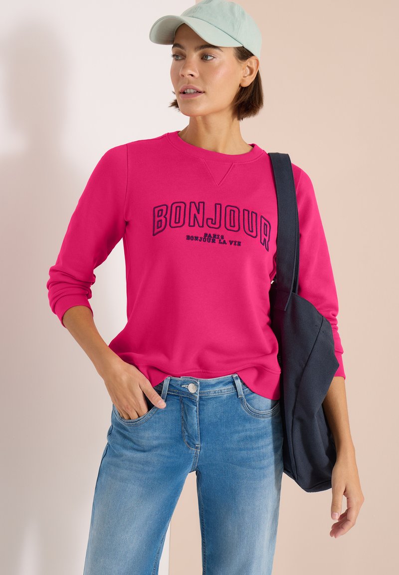 Femme portant une casquette vert clair, un sweat-shirt rose vif avec le texte « BONJOUR », un jean bleu et un sac à bandoulière noir sur un fond neutre.