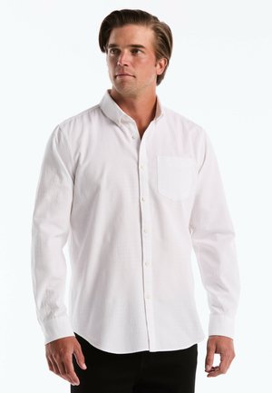 Man draagt een wit overhemd met lange mouwen en een borstzak, staat en kijkt iets opzij.