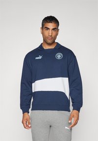 Pulover navy cu dungi orizontale albe; are un guler, logo Puma și emblema Manchester City pe piept. Blană moale din amestec de bumbac.