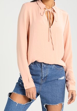 Vrouw die een lichtroze blouse met lange mouwen draagt, met een gestrikt bovenstuk en versleten blauwe spijkerbroek met grote scheuren op de dijen.