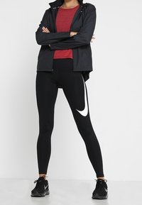 Leggings de sport noires avec un swoosh blanc de Nike, associés à un haut texturé rouge et une veste noire zippée. Des chaussures de course noires complètent le look.