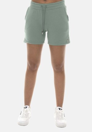 Personne portant un short vert sauge avec cordon et des baskets blanches, debout les mains dans les poches sur un fond blanc uni.