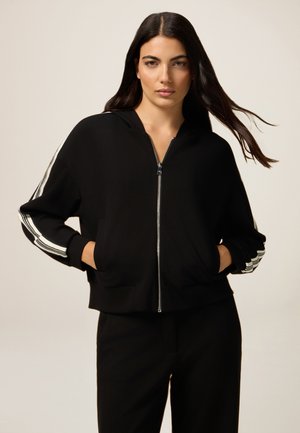 Black zip-up hoodie with a fitted design, large front pockets, and three white stripes along the sleeves. Smooth fabric, relaxed fit.
