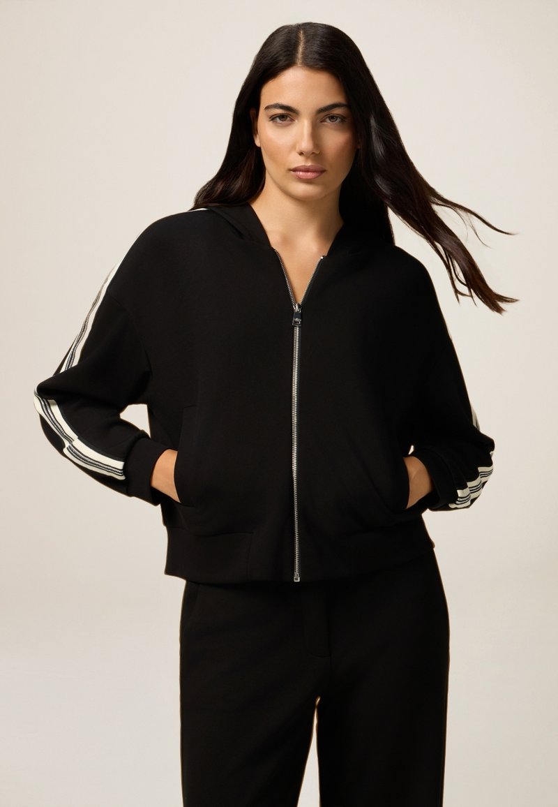 Zwarte rits hoodie met een getailleerd ontwerp, grote zijzakken en drie witte strepen langs de mouwen. Glad materiaal, ontspannen pasvorm.
