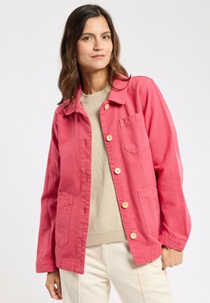 Femme aux longs cheveux bruns portant une veste rouge à boutons sur un pull beige et un pantalon clair, debout devant un fond uni.