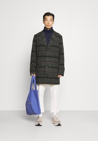 Grå rutete frakk med grønne striper, enkel spent design, brukt over en marineblå turtleneck. Modellen holder en blå tote bag og har på seg lyse sneakers.