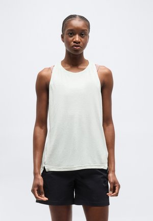 Femme aux cheveux tressés portant un haut blanc sans manches et un short noir, debout devant un fond clair uni.