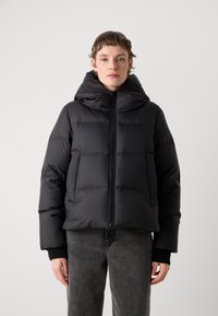 Doudoune noire avec capuche, dotée d'une fermeture éclair et de larges coutures horizontales, associée à un pantalon en denim gris.