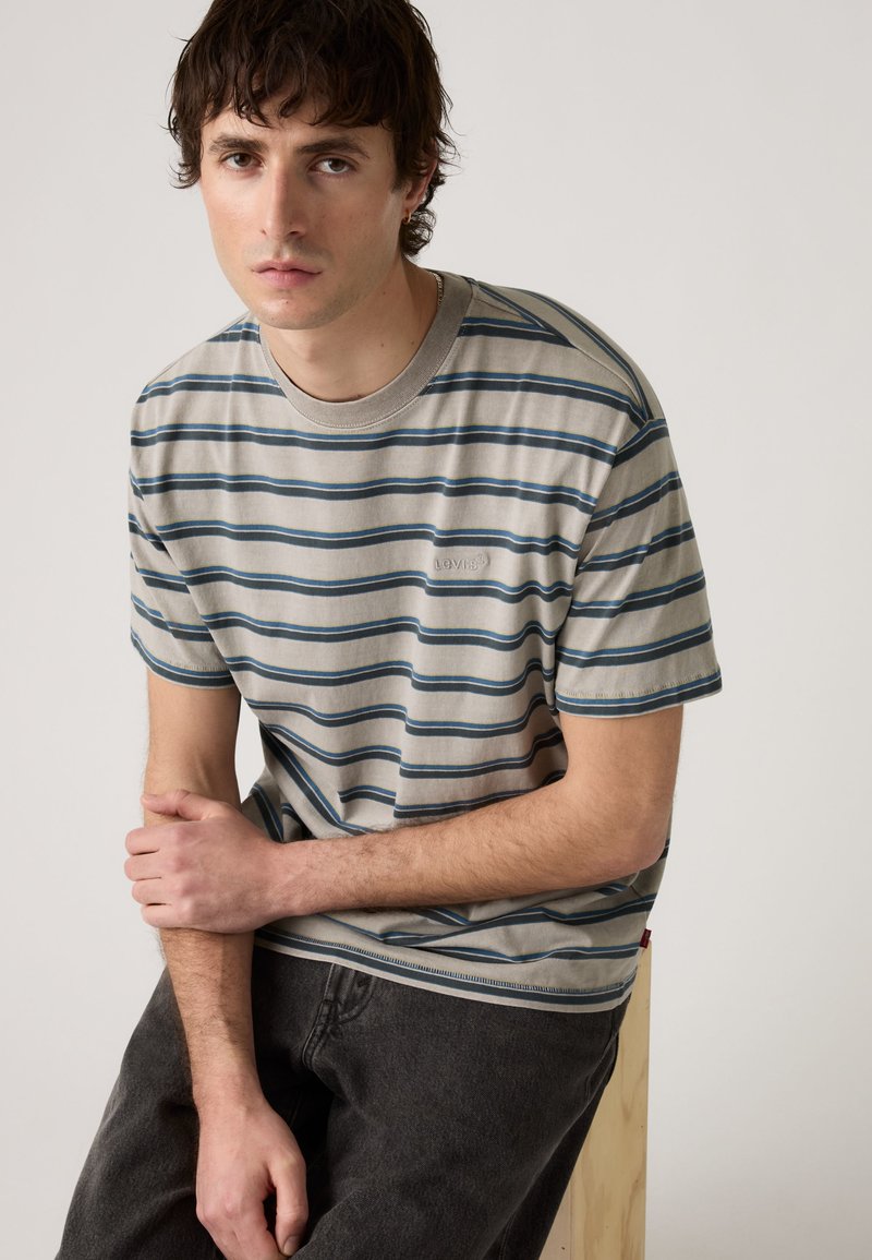 Young man sitting, wearing a beige and blue striped Levi's t-shirt and dark jeans, with one arm resting on the other.