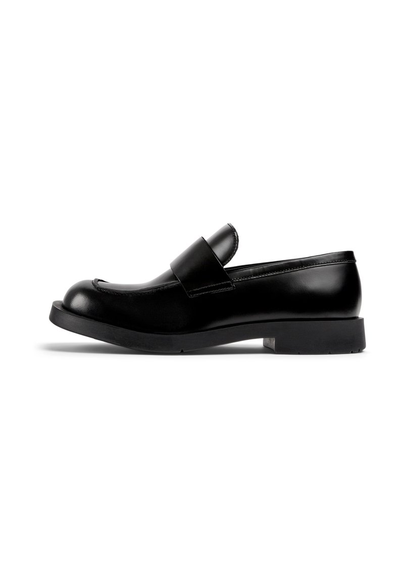 Mocassin en cuir noir avec un bout arrondi, semelle en caoutchouc plate et un détail de bandeau proéminent sur le devant. Texture lisse, design minimaliste.