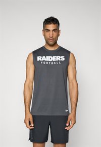 Siva brezrokavnik s tekstom "RAIDERS Football" v belem. Narejen iz gladkega materiala. V par z temnimi hlačami, na katerih je ob strani napis "RAIDERS".