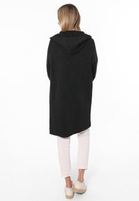 Schwarze, langärmelige Kapuzenjacke mit weicher Textur, geradem Schnitt und überschnittenen Schultern. Kombiniert mit hellrosanen Hosen und beigen Schuhen.
