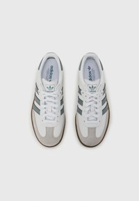 Biele tenisky s šedými semišovými akcentmi a tromi tmavosivými pruhmi. Majú textúrovanú gumovú podrážku a biele šnúrky. Viditeľné logo Adidas.