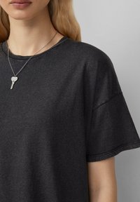 Zwarte T-shirt met ronde hals, gemaakt van zacht materiaal en met korte mouwen. Een zilveren ketting met een hartvormige sleutelhanger is ook zichtbaar.