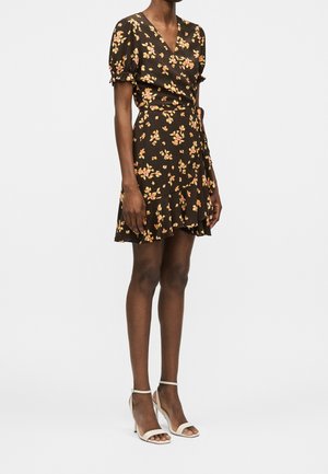 Vrouw draagt een zwarte wikkeljurk met roze en gele bloemenprint, gecombineerd met witte sandalen met hoge hakken, staand tegen een effen achtergrond.