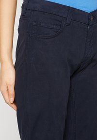 Tummansiniset chinot, joissa on sileä pinta, varustettu tavallisilla etutaskuilla, koristeommella sekä nappikiinnityksellä vyötäröllä.