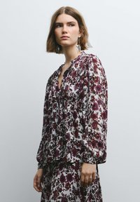 Blusa floreale in tessuto bianco con fiori bordeaux e viola scuro, maniche lunghe a sbuffo e scollo a v.