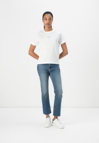 White cotton t-shirt with short sleeves and a round neckline, featuring blue text on the chest. Paired with blue denim jeans and white sneakers.
