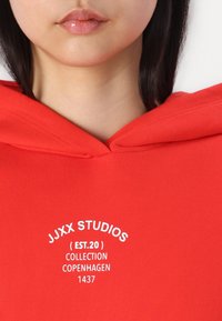 Piros kapucnis pulóver bordázott textúrával, fehér ívelt logóval: "JJXX STUDIOS (EST.20) KOLEKCIÓ KOPENHÁGA 1437."