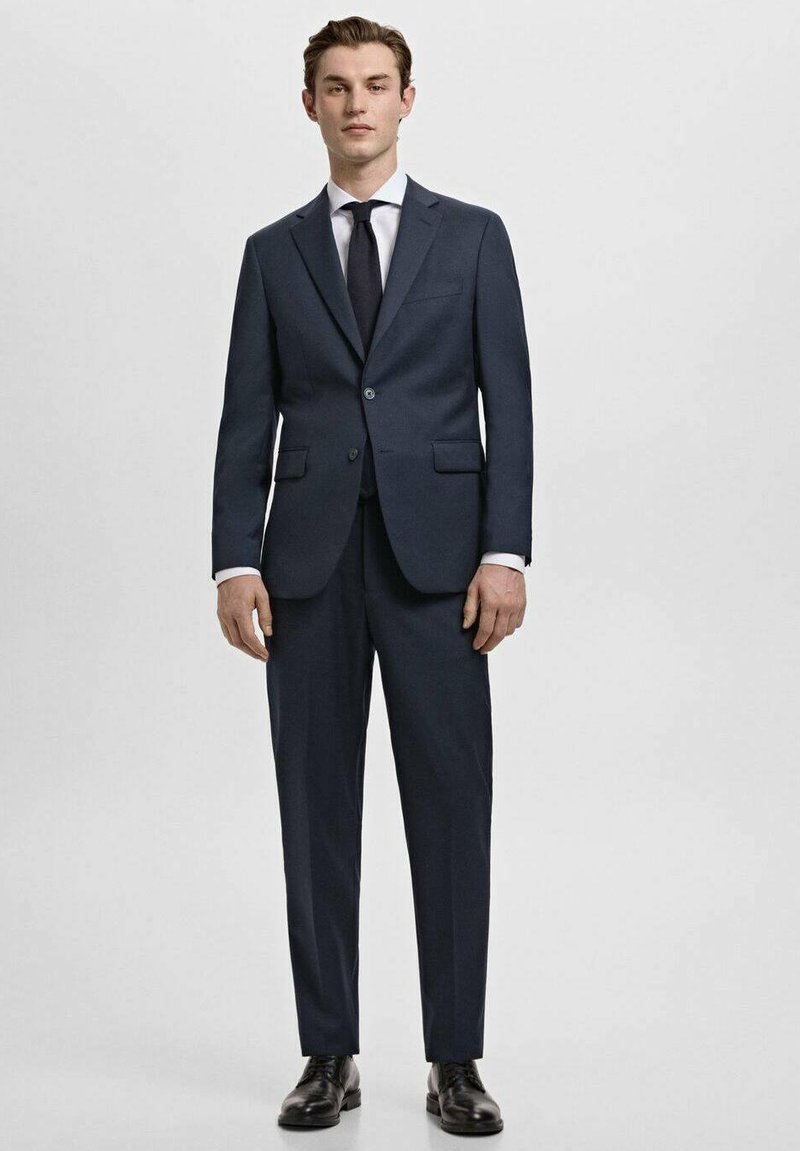 Costume bleu marine avec une veste à deux boutons et un pantalon droit. Chemise blanche et cravate sombre complètent le look, assortis à des chaussures noires.