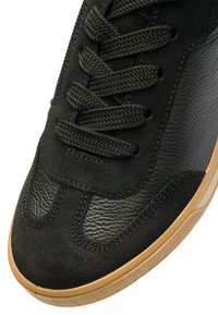 Black sneaker featuring suede and textured leather upper, with flat black laces and a gum rubber sole. Rounded toe and stitched accents.