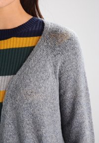 Grå stickad cardigan med en mjuk textur, med V-hals och en lös passform, bärs över en flerfärgad randig tröja i marinblått, gult och grönt.