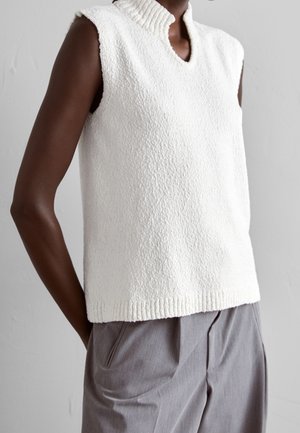 Bezrekawnik z białego dzianinowego materiału z fakturą, z prążkowanym kołnierzem i wykończeniem na dole, noszony z luźnymi szarymi spodniami na jednolitym tle.