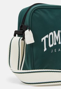 Zelená nylonová taška s černým zipem. Má nastavitelný popruh s bílými pruhy a výrazný bílý text "TOMMY JEANS" na jedné straně.