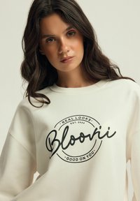 Kremowa bluza z okrągłym logo z napisem "Bloori" oraz tekstem "REAL LOOKS. EST. 2024. GOOD ON YOU" w kolorze czarnym. Miękki materiał i luźny krój.