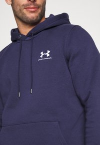 Marine hoodie van een zachte stof met een kangoeroezak, voorzien van een wit Under Armour-logo op de borst en trekkoorden voor aanpassing.