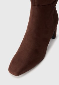 Καφέ σουέτ μποτάκι μέχρι τον αστράγαλο με τετράγωνη μύτη, κοντό κυλινδρικό τακούνι και λεία υφή. Ο σχεδιασμός είναι μινιμαλιστικός, χωρίς ορατές διακοσμήσεις.